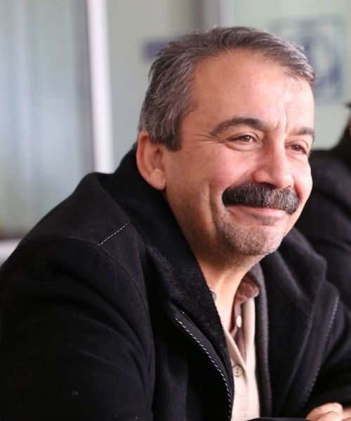 Sırrı Süreyya Önder. Kapalı bir mekânda oturuyor ve gülümserken görülüyor. Koyu renk bir mont ve açık renk bir gömlek giymiş. Arka planda bulanık biçimde masa ve duvar detayları seçiliyor. Işık, kişinin yüzünü yumuşak bir şekilde aydınlatıyor.