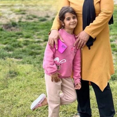 On a light green grassy patch, Narin, wearing a pink sweater with a rabbit pattern and light beige pants, leans against her mother Yüksel Güran’s arm and smiles. Yüksel Güran’s face is out of frame; she is seen wearing a mustard-yellow tunic, navy pants, and a black headscarf. Shot in natural light in a simple rural setting, the image reflects the closeness and warmth between mother and daughter.